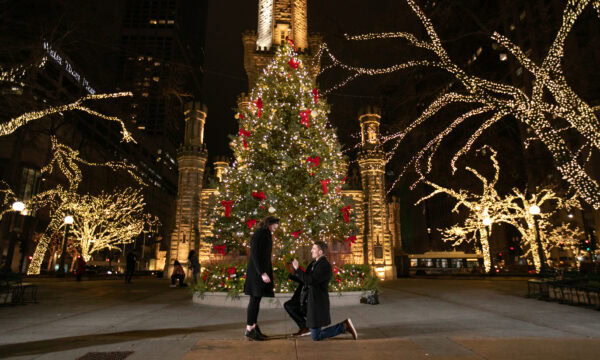 a man is down on one knee asking his girlfriend to marry him in front of a christmas tree at night at the chicago pumping station