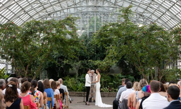 two brides stand in front of their guests as they exchange rings during their wedding ceremony in a greenhouse